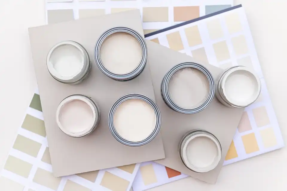 Selección de pinturas de pared: seis latas de pintura abiertas en tonos de blanco y crema dispuestas en diagonal sobre muestras de color.