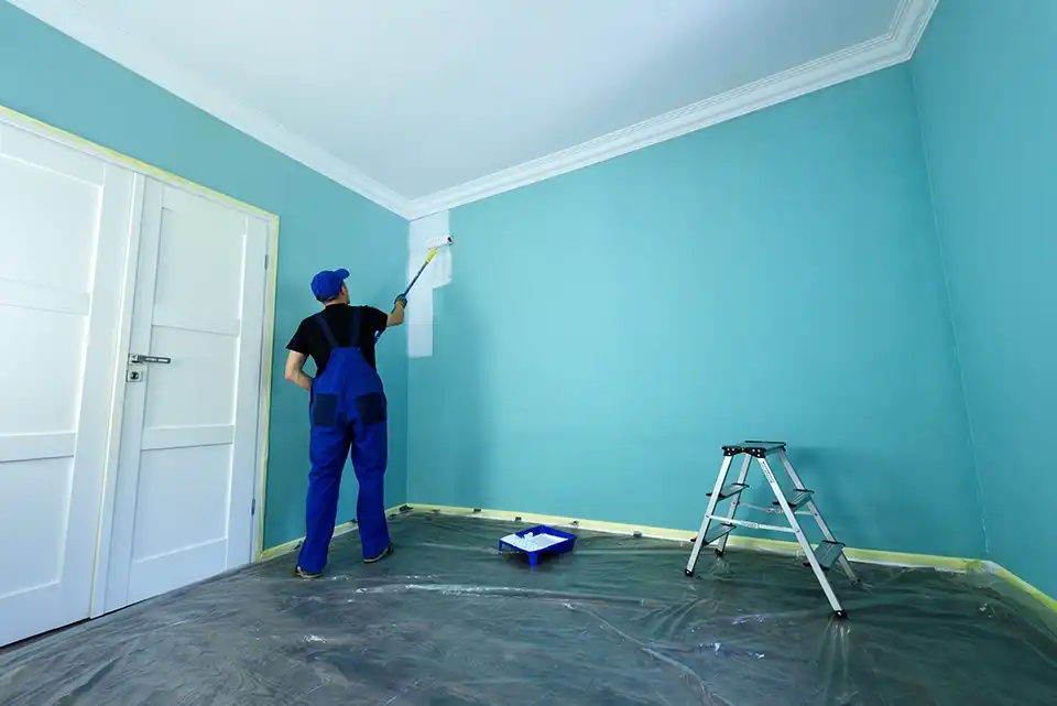 Un pintor con mono de trabajo azul pintando con rodillo una pared interior de color turquesa, con una escalera y bandeja de pintura a su lado, en una habitación protegida con lona en el suelo.