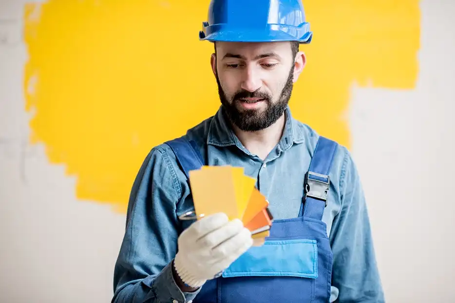 Un pintor con casco de seguridad azul y overol mirando muestras de color naranja con una pared parcialmente pintada de amarillo en el fondo.