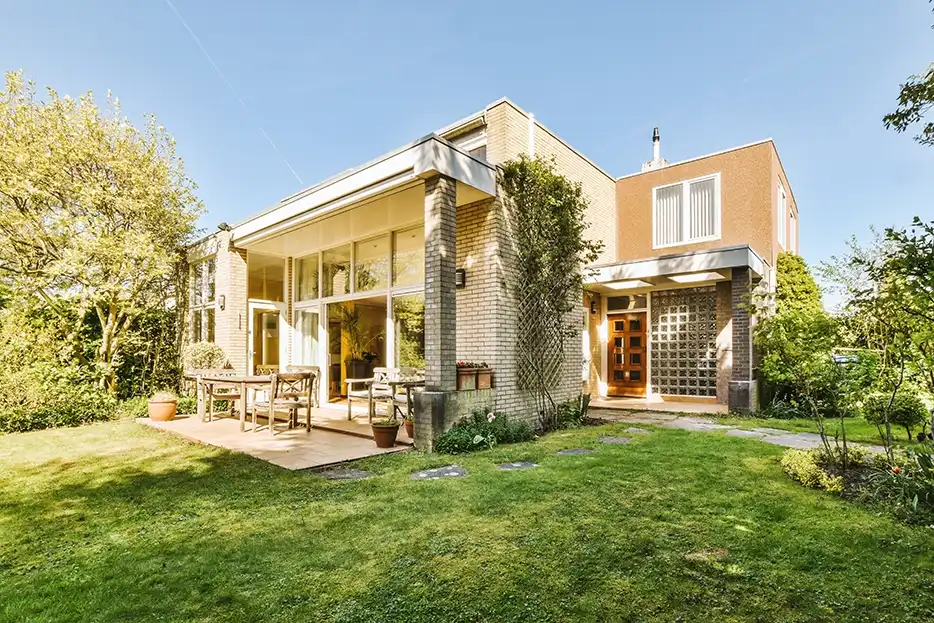 Vista panorámica de una casa de ladrillos con grandes ventanas y una terraza con mobiliario de madera. El jardín al frente está bien cuidado, con césped verde, árboles y plantas.