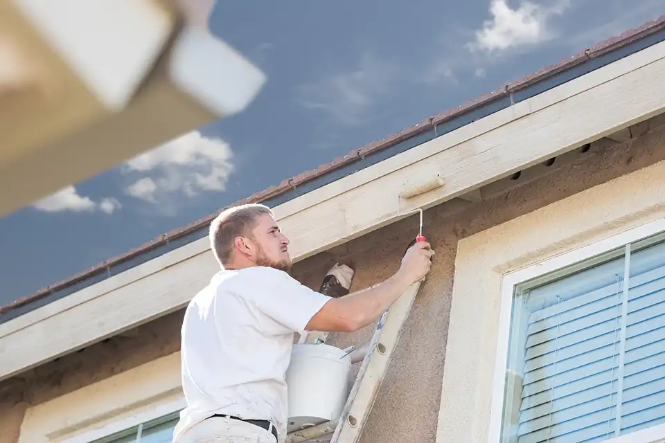 Un pintor profesional en una escalera utilizando un rodillo pequeño para pintar el acabado exterior de una casa residencial de color beige bajo un cielo azul despejado.