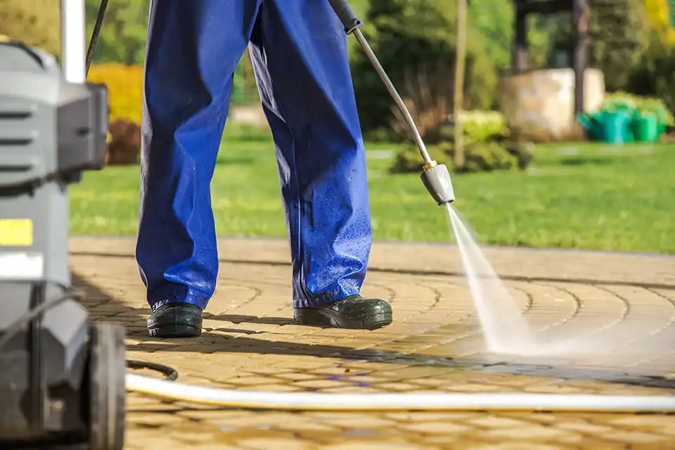 Persona con mono de trabajo azul utilizando una hidrolavadora para limpiar un pavimento estampado en un jardín.